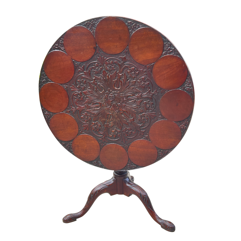 A round wooden table with intricate carvings around the edge, featuring a floral motif in the center, with a simple triangular pedestal base.