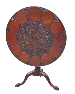 A round wooden table with intricate carvings around the edge, featuring a floral motif in the center, with a simple triangular pedestal base.